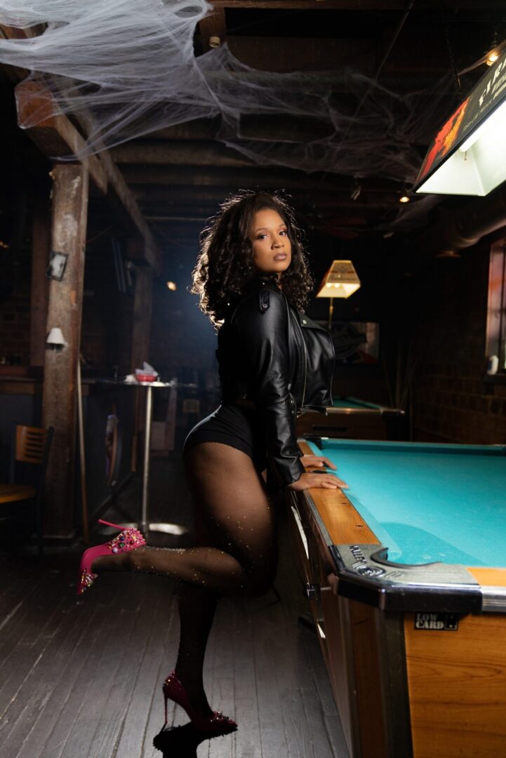 A woman in black leather jacket standing next to pool table.